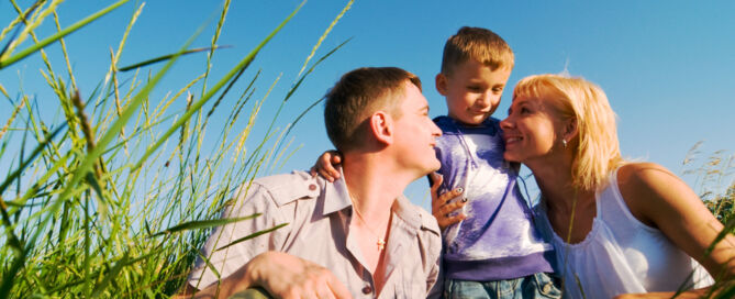 A low-angle shot shows a family of three—a man, a woman, and a young boy—in a field of tall green grass under a clear blue sky.