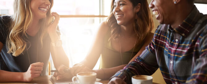 'What is Naloxone' featured image - Three young adults, two women and a man, are seated at a wooden table in a cafe, laughing and talking over coffee.