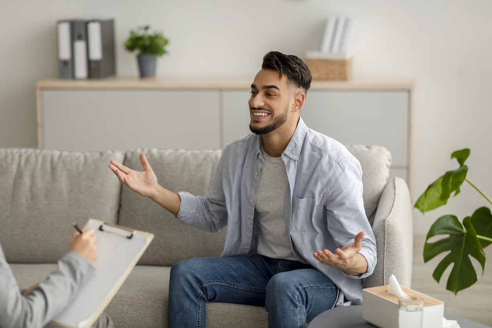 A man smiles and gestures during individual therapy, possibly for opioid addiction, while someone takes notes.